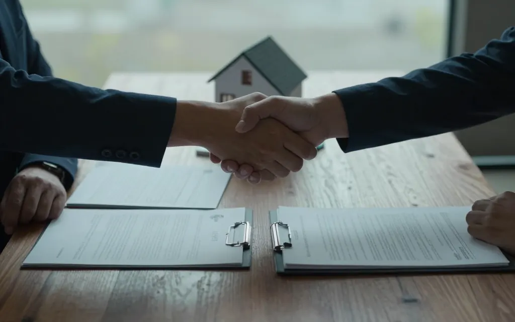 Dos personas se dan la mano sobre una mesa, con un contrato firmado y una maqueta de casa entre ellas.